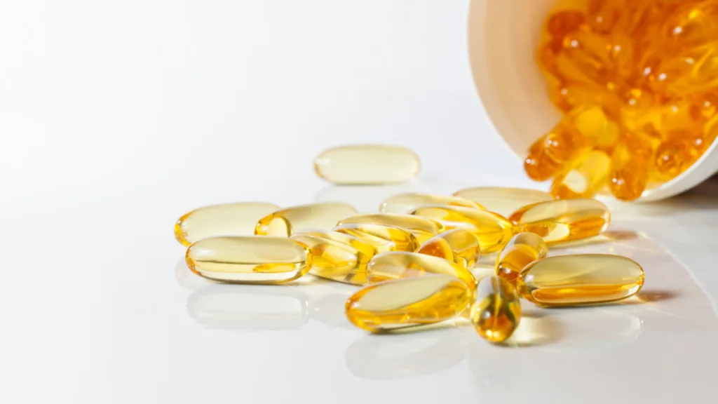 Close-up of yellow gel capsule supplements spilling out of a white container onto a reflective surface.