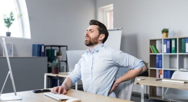 Man sitting at desk holding his lower back in discomfort while working at a computer.
