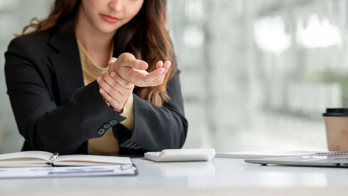 Office worker holding her wrist in pain while sitting at a desk, illustrating a repetitive stress injury such as carpal tunnel syndrome caused by prolonged computer and desk work.