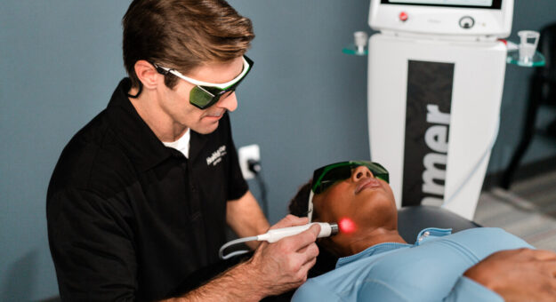 Chiropractor administering Class IV laser therapy to a patient's neck while both wear protective eyewear in a clinical treatment room. A red laser light is visible at the treatment site as the patient lies comfortably on a treatment table.