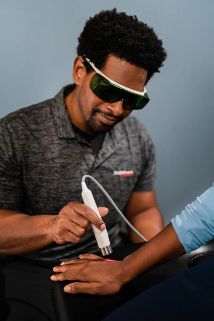 Healthcare provider wearing protective laser safety glasses while performing Class IV laser therapy on a patient's hand. A red treatment light is visible on the patient's wrist as they receive care in a clinical setting.